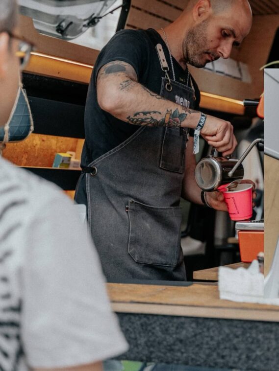 A barista with tattoos serving coffee from a mobile coffee van